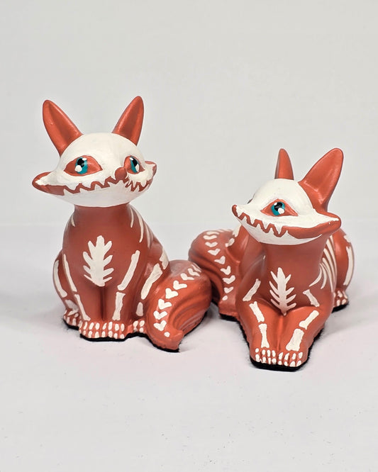 Two skeleton fox figurines with white faces and terracotta orange bodies on a light gray background